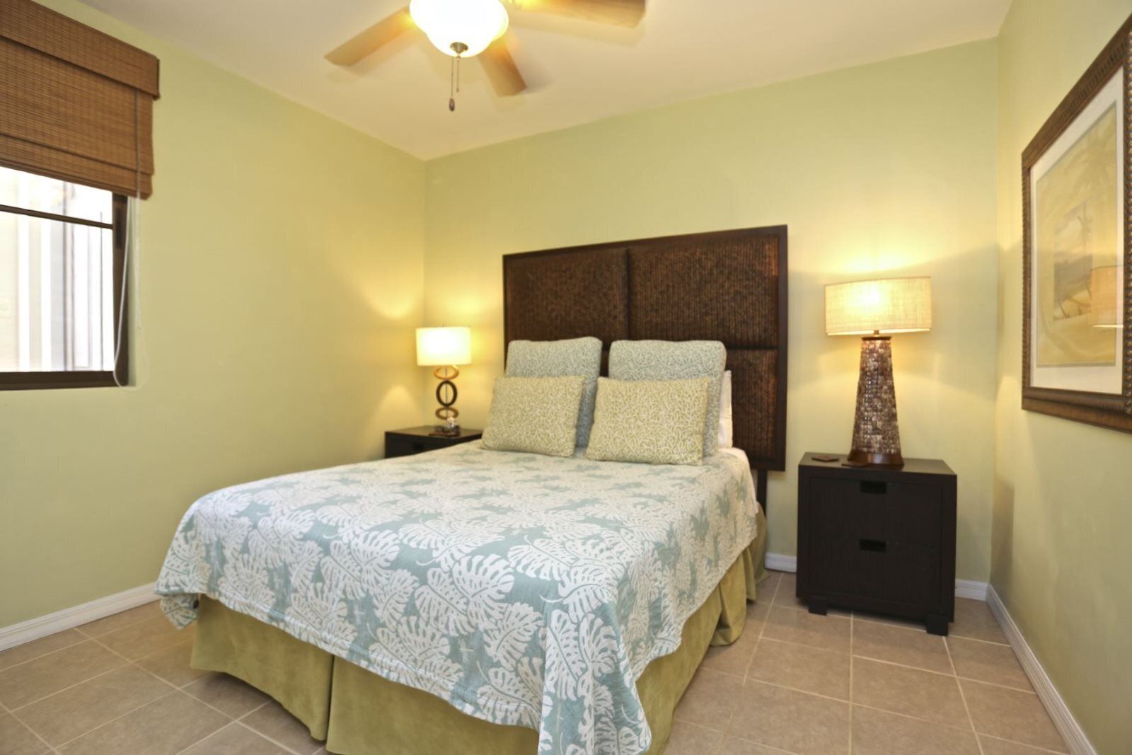 Une chambre à coucher comprenant un lit, deux tables de chevet, un ventilateur de plafond et une fenêtre avec des stores.