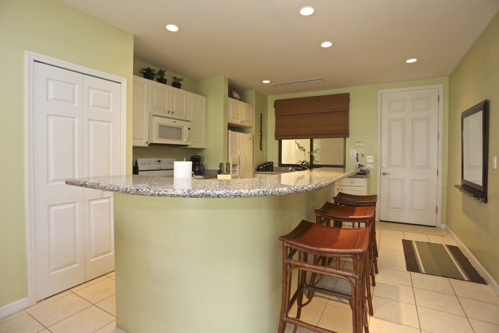 A kitchen with a granite countertop, white cabinets, a microwave, and a window with blinds. There are three stools and a door with a mirror.
