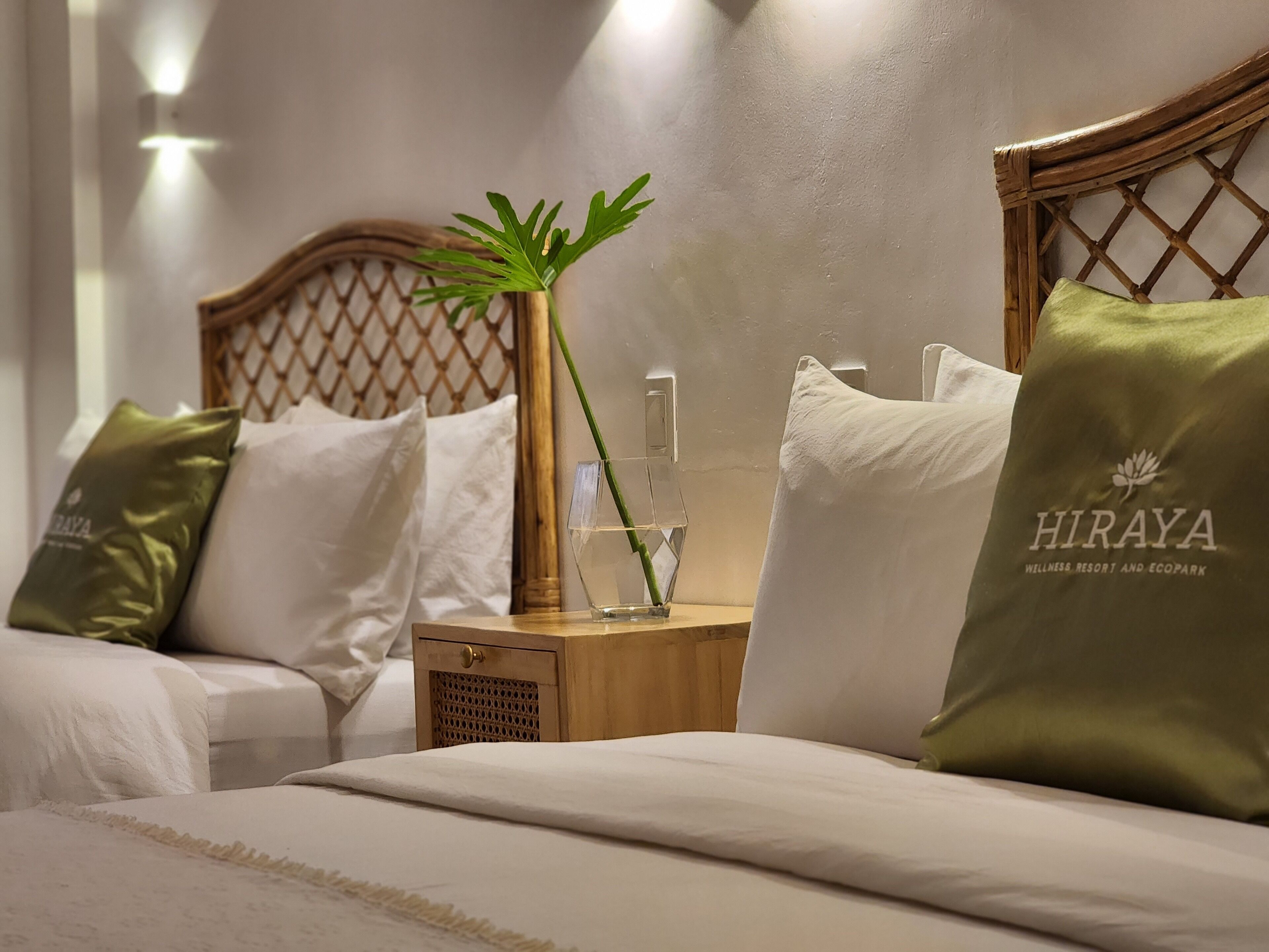 A hotel room with two beds, each with a pillow labeled "HIRAYA". A wooden nightstand with a glass vase and a bottle is between the beds.