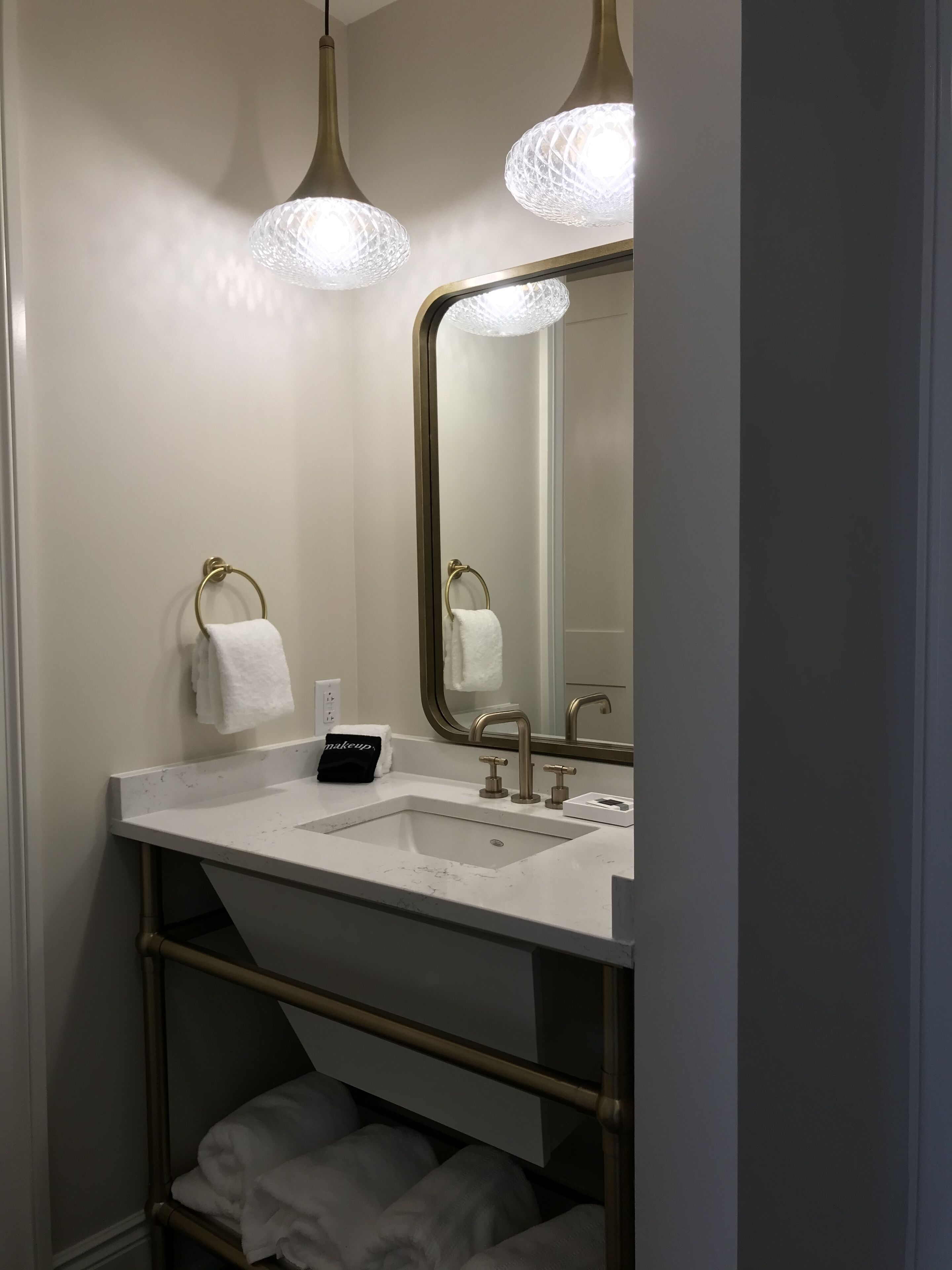Un baño con un lavamanos blanco, dos argollas doradas para toallas, un espejo con marco dorado y un perchero.