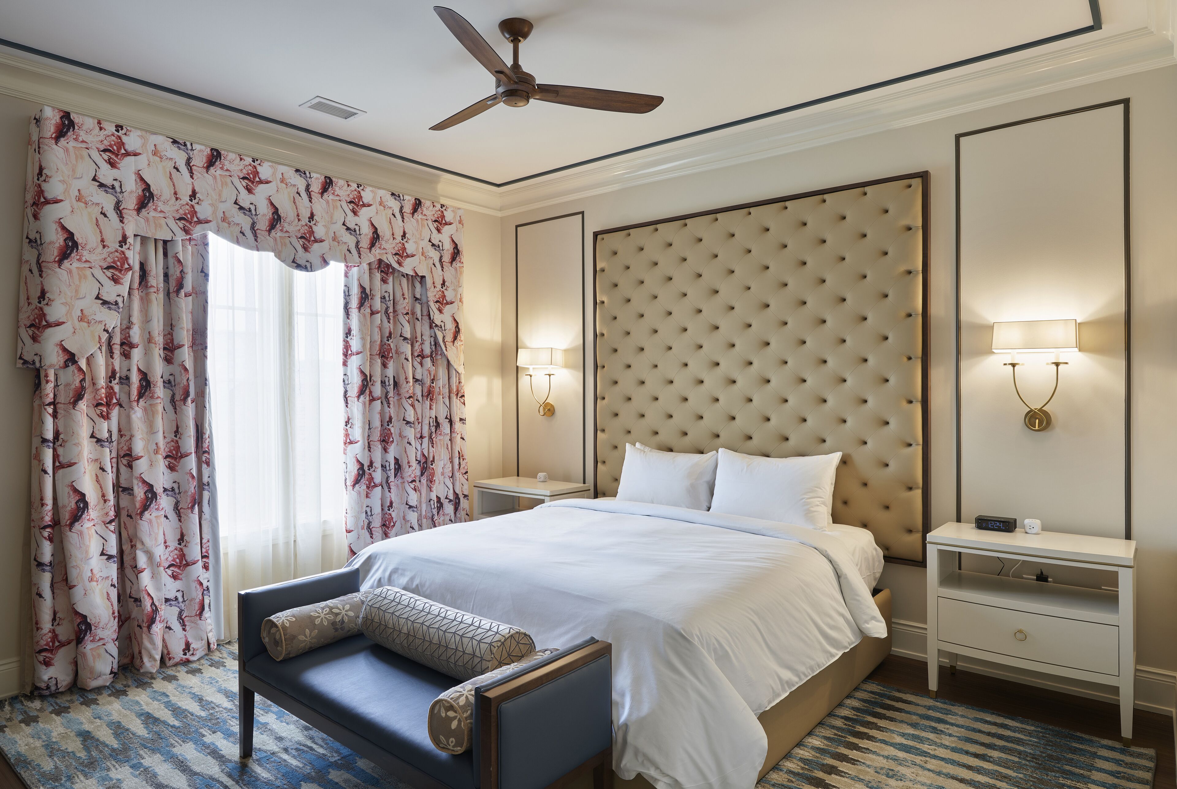 A hotel room with a large bed, a ceiling fan, a nightstand with a lamp, and a window with floral curtains.