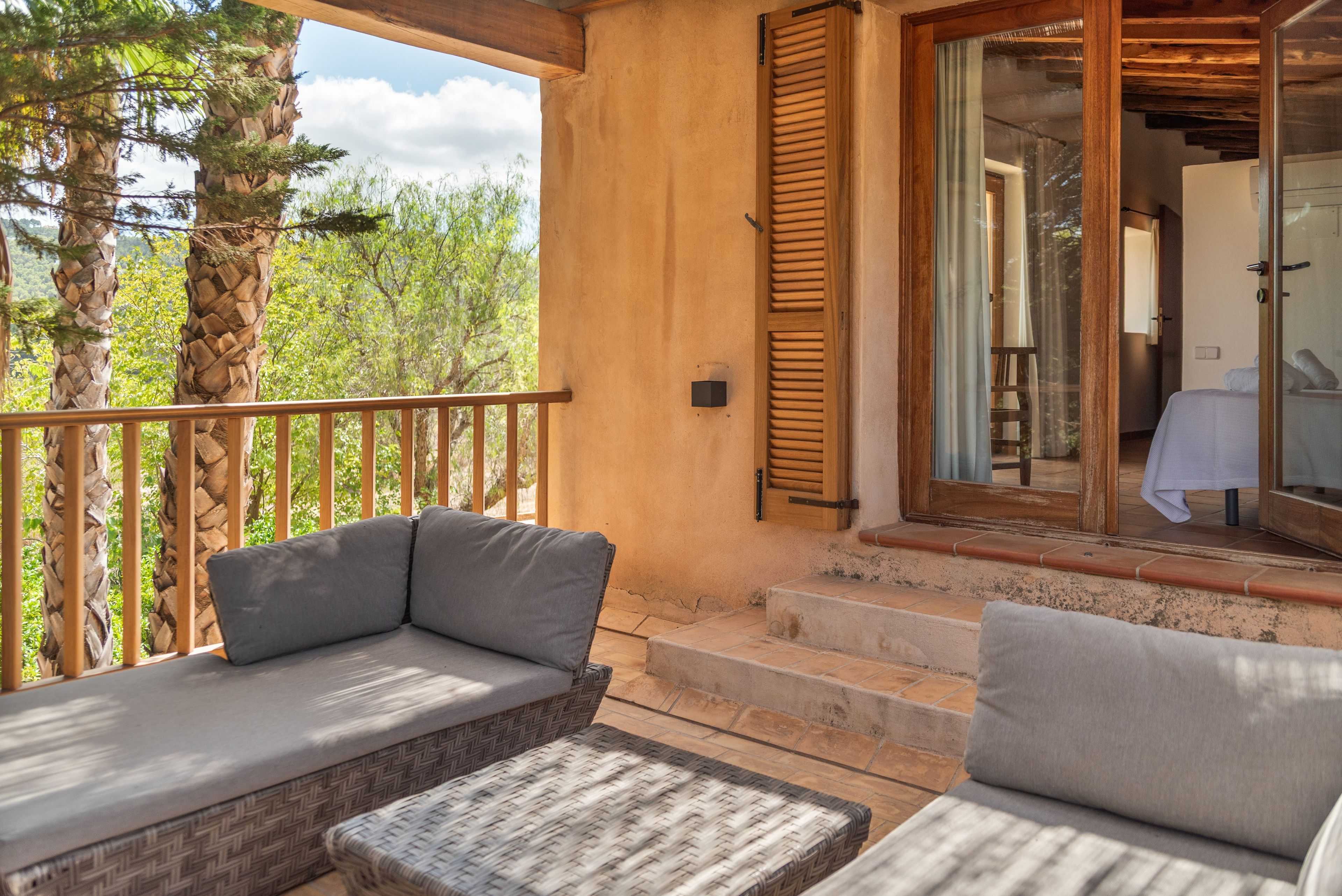 Un balcon avec un canapé et un pouf en osier, une porte en bois donnant sur une vue agréable et un mur en pierre.