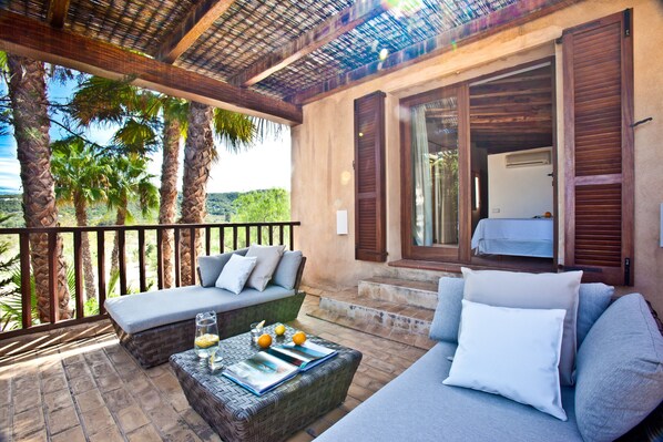 A balcony with a sofa, a wicker coffee table with magazines and fruit, and a view of palm trees and hills.
