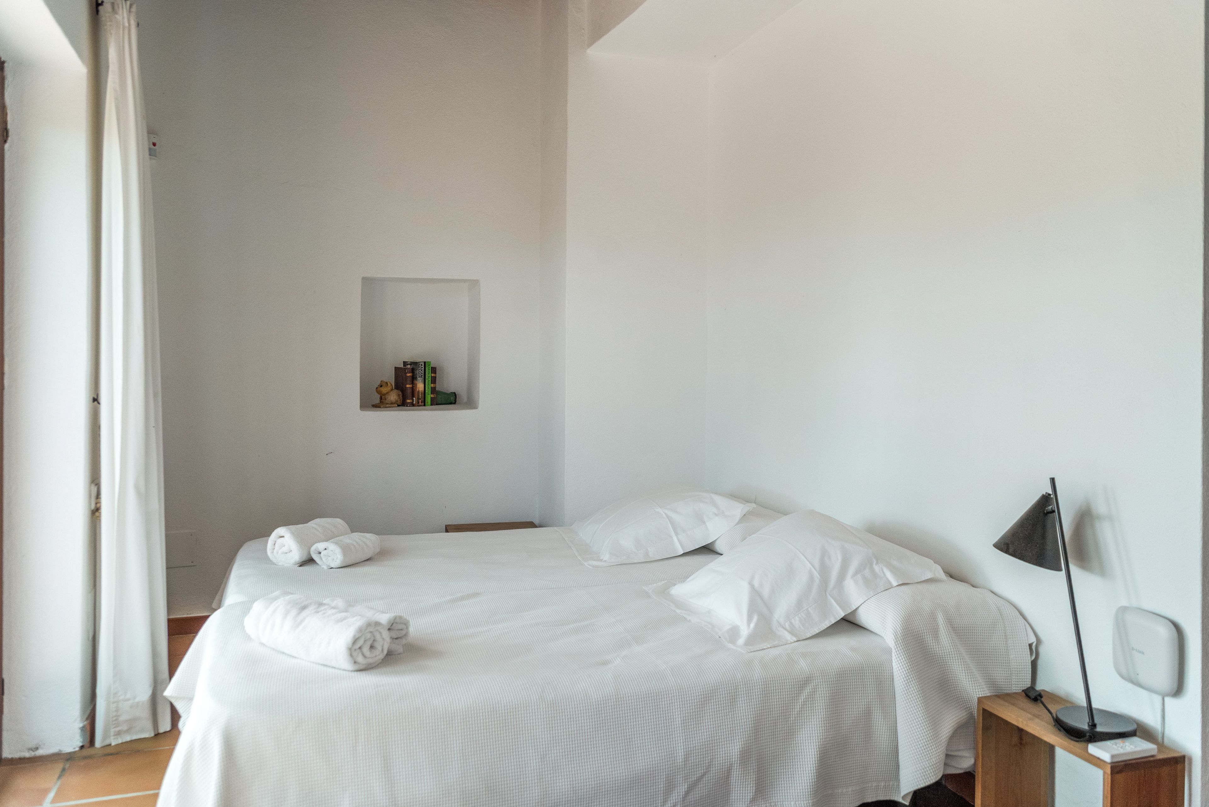Ein ordentlich gemachtes Bett mit weißer Bettwäsche, ein Nachttisch mit Lampe und ein kleines Regal mit Büchern und einem Teddybär.