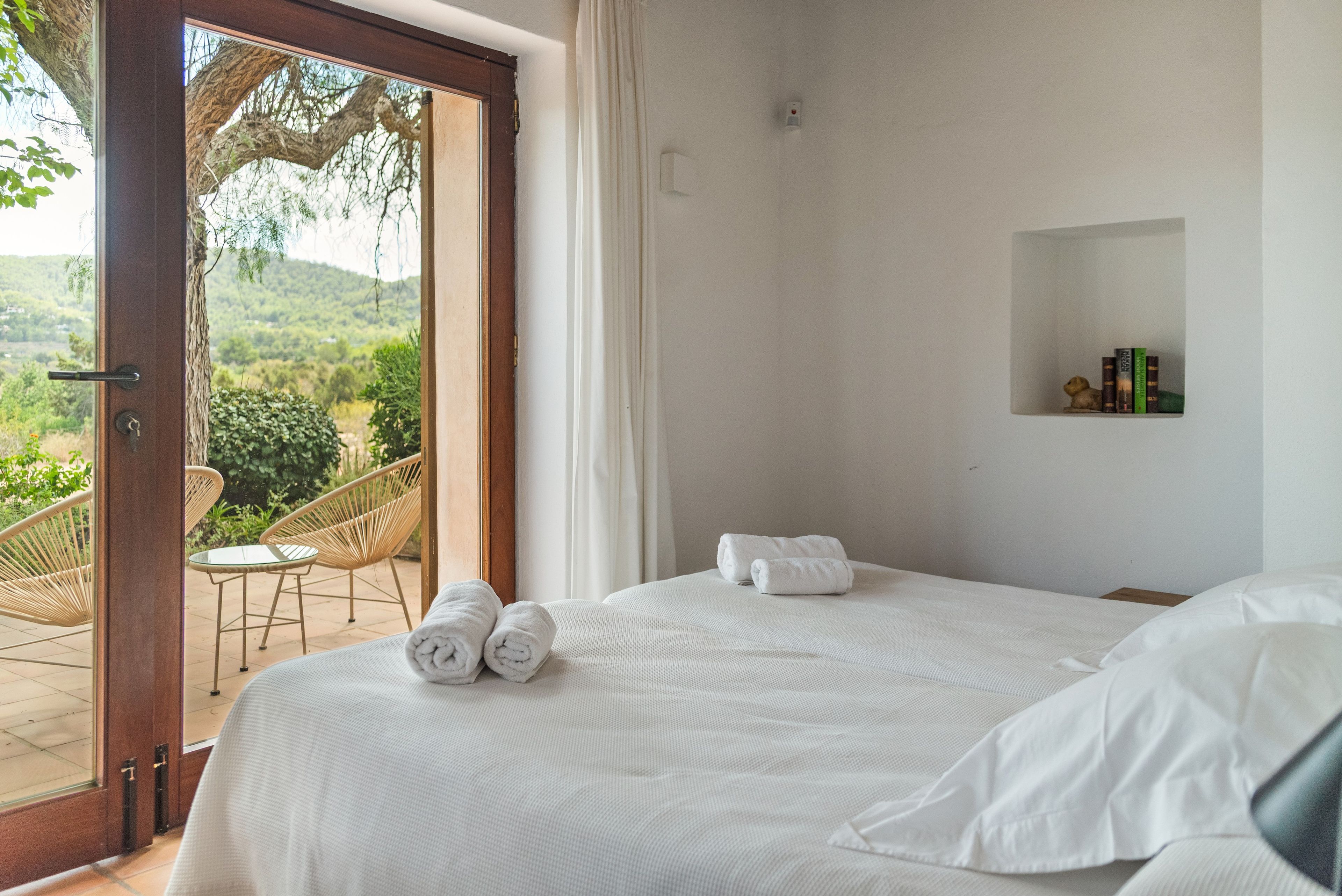 A bedroom with a large bed, two rolled towels, a small shelf with books, and a view of a garden through an open door.