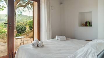 A bedroom with a large bed, two rolled towels, a small shelf with books, and a view of a garden through an open door.