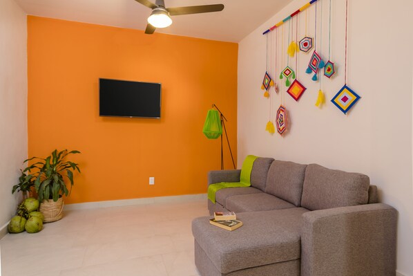 A living room with a grey sofa, a flat-screen TV mounted on an orange wall, a ceiling fan, a hanging decorative piece, and a potted plant. - Riviera Sayulita Hotel (Sayulita)