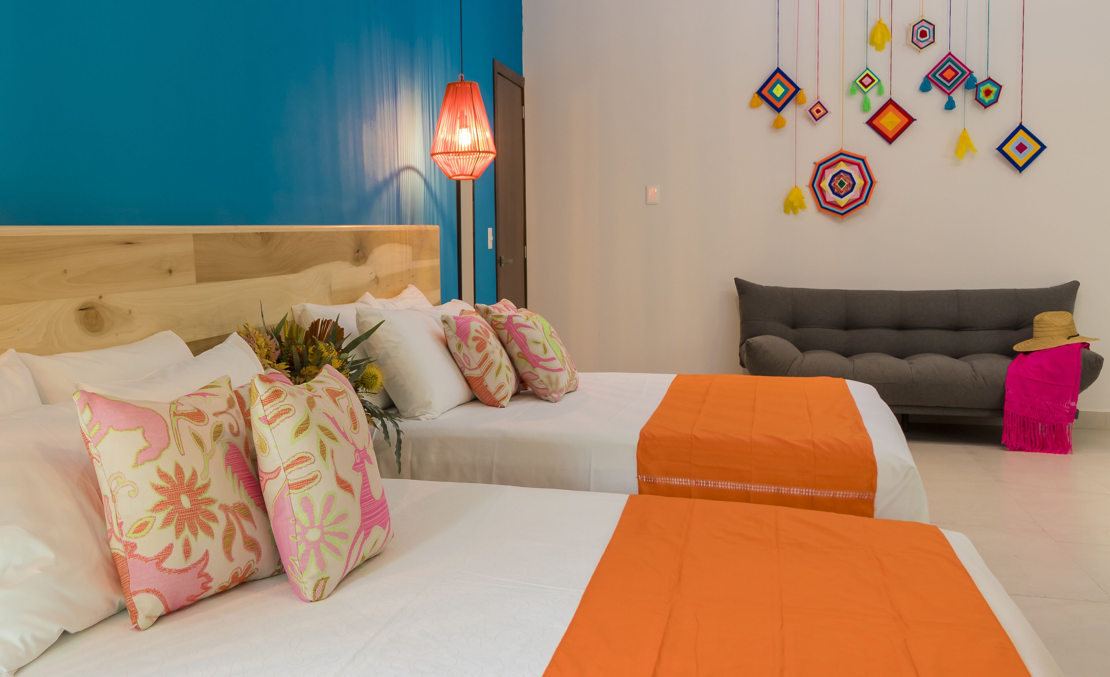 A hotel room with two beds, a grey sofa, a colorful wall decoration, and a hanging lamp.