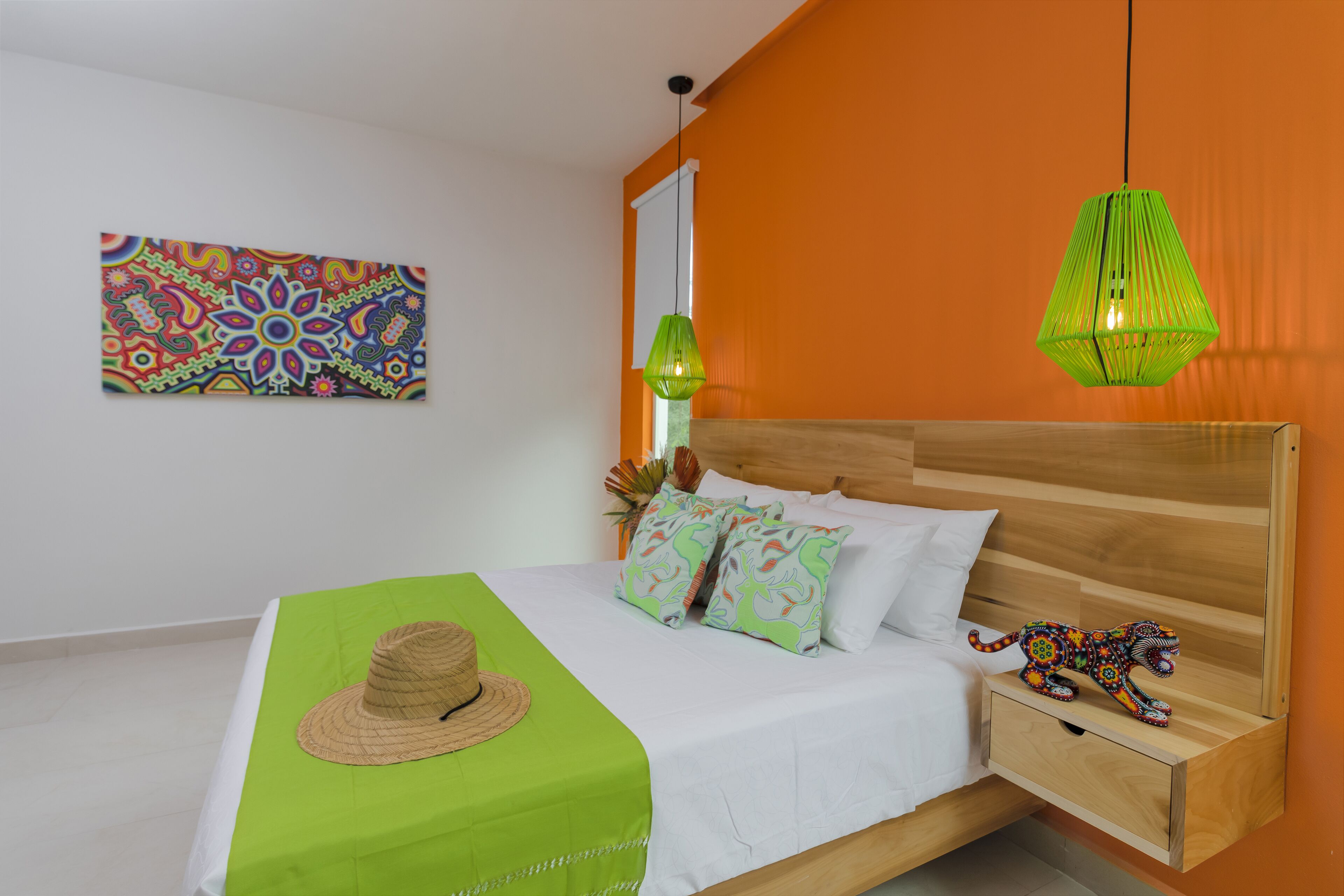 Un dormitorio con cama de madera, colcha verde, almohadas decorativas, un sombrero sobre la cama, una obra de arte colorida en la pared y una lámpara colgante verde.