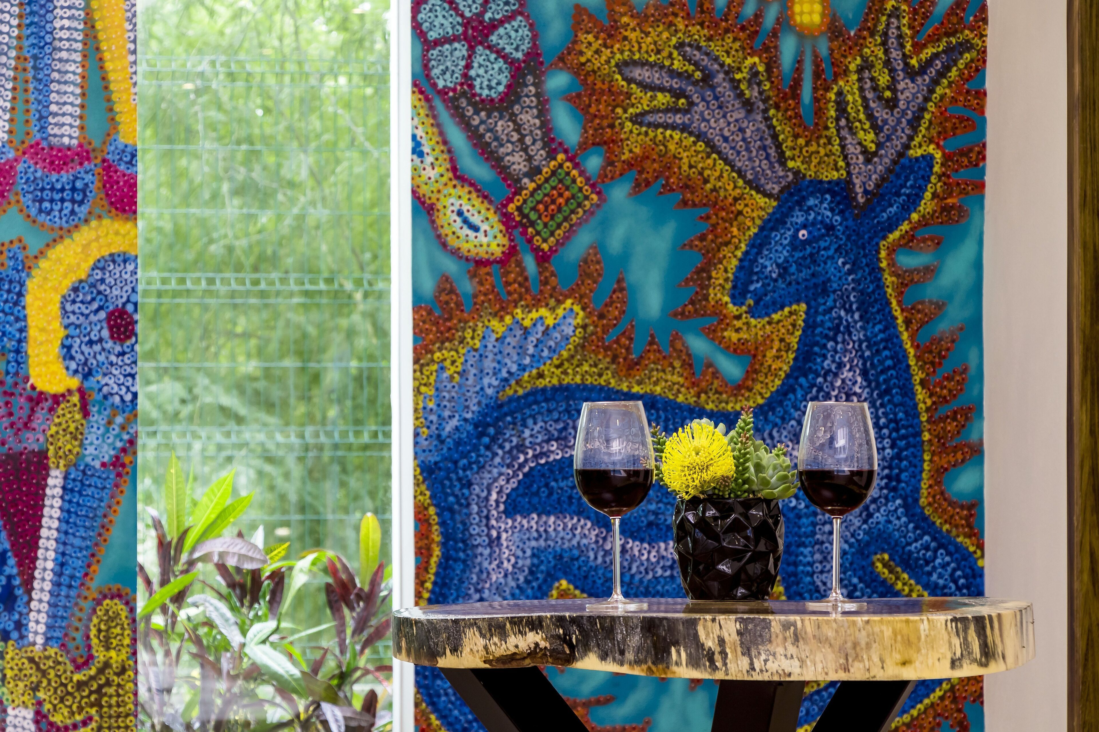 A table with two glasses of red wine and a vase with yellow flowers against a colorful tapestry backdrop.
