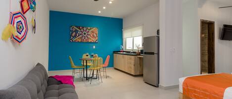 A modern room with a grey sofa, a dining area with yellow chairs, a kitchen with a fridge, and a bed with an orange blanket.