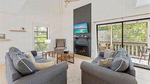 Een ruime woonkamer met grijze banken, een houten salontafel, een open haard en een aan de muur bevestigde televisie.