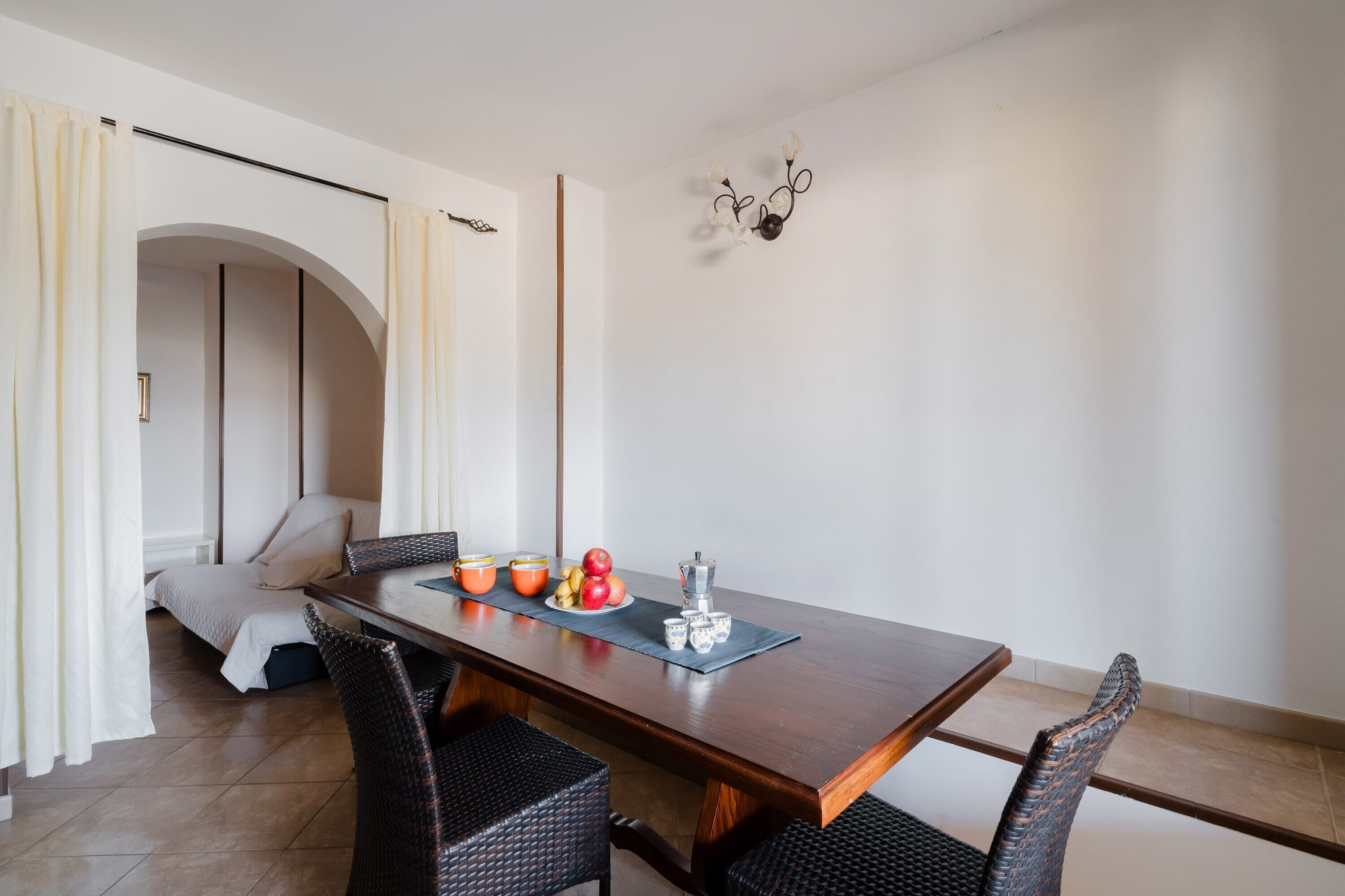 A dining area with a wooden table, black wicker chairs, a chandelier, and a bed in the background.