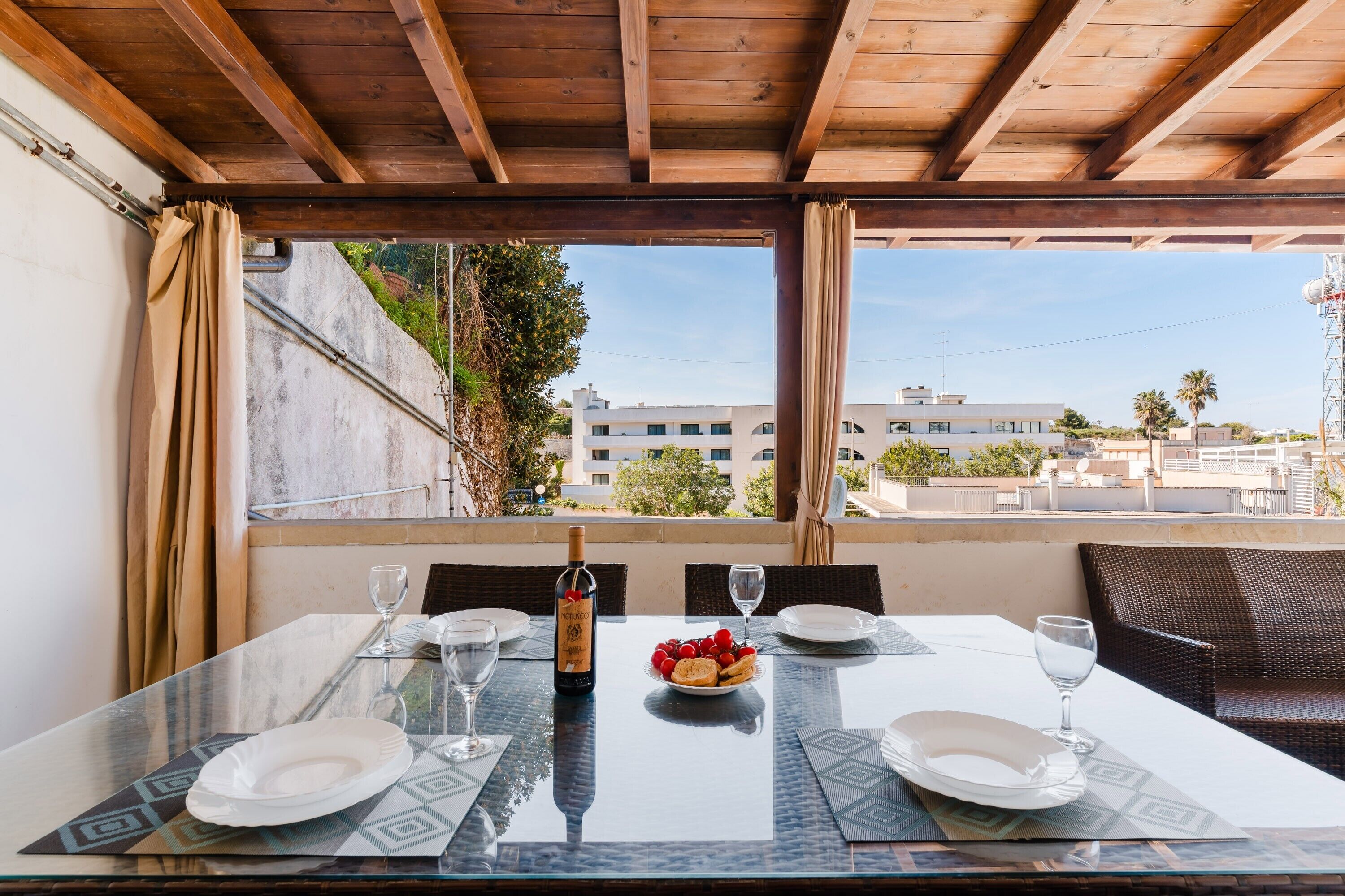 A dining table with a bottle of wine, glasses, and a plate of fruit in a room with a wooden ceiling and large windows.