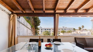 Une table à manger avec une bouteille de vin, des verres et une assiette de fruits, dans une pièce dotée d’un plafond en bois et de grandes fenêtres.