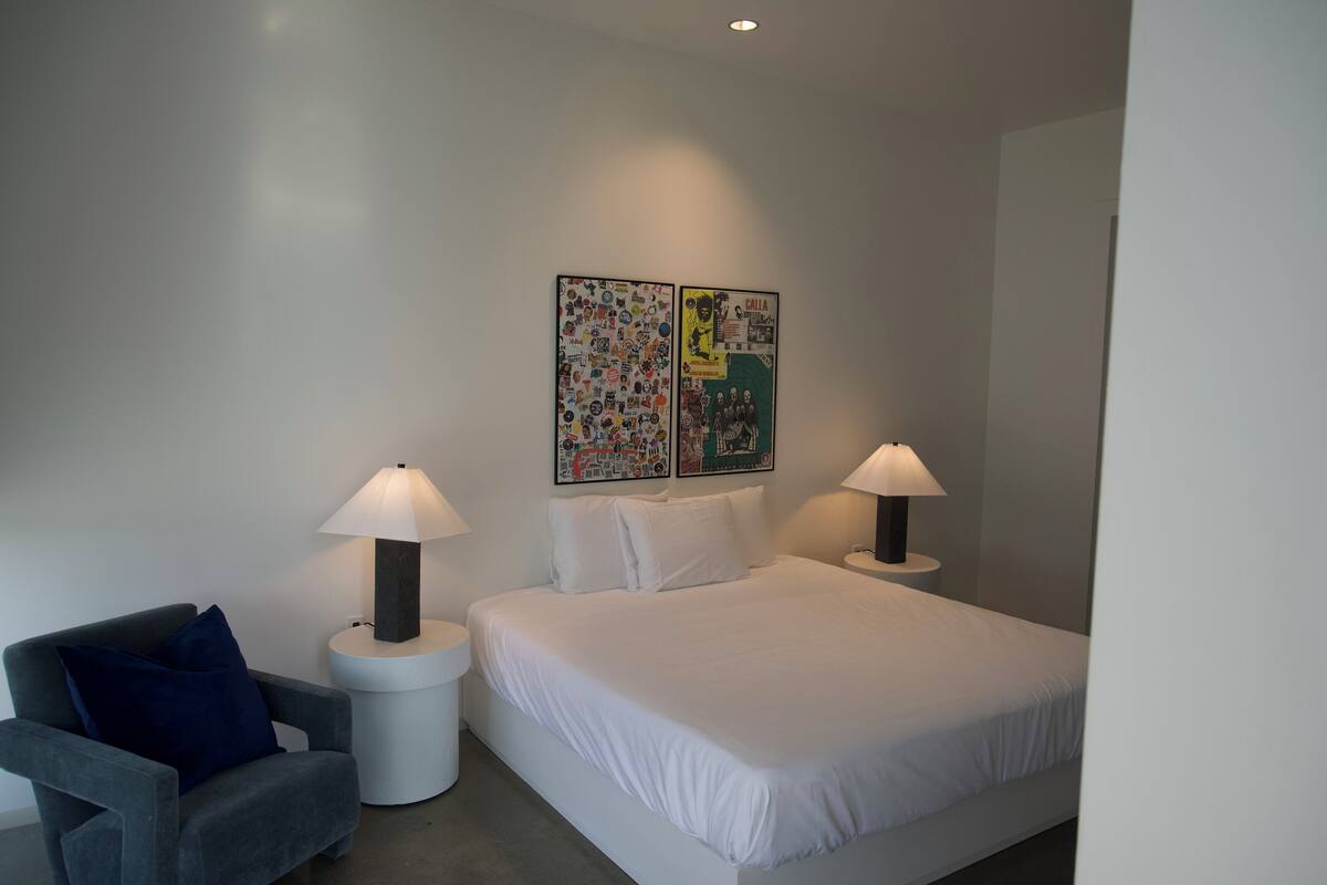 A modern bedroom with a large bed, two bedside tables with lamps, a grey armchair, and two wall art pieces.