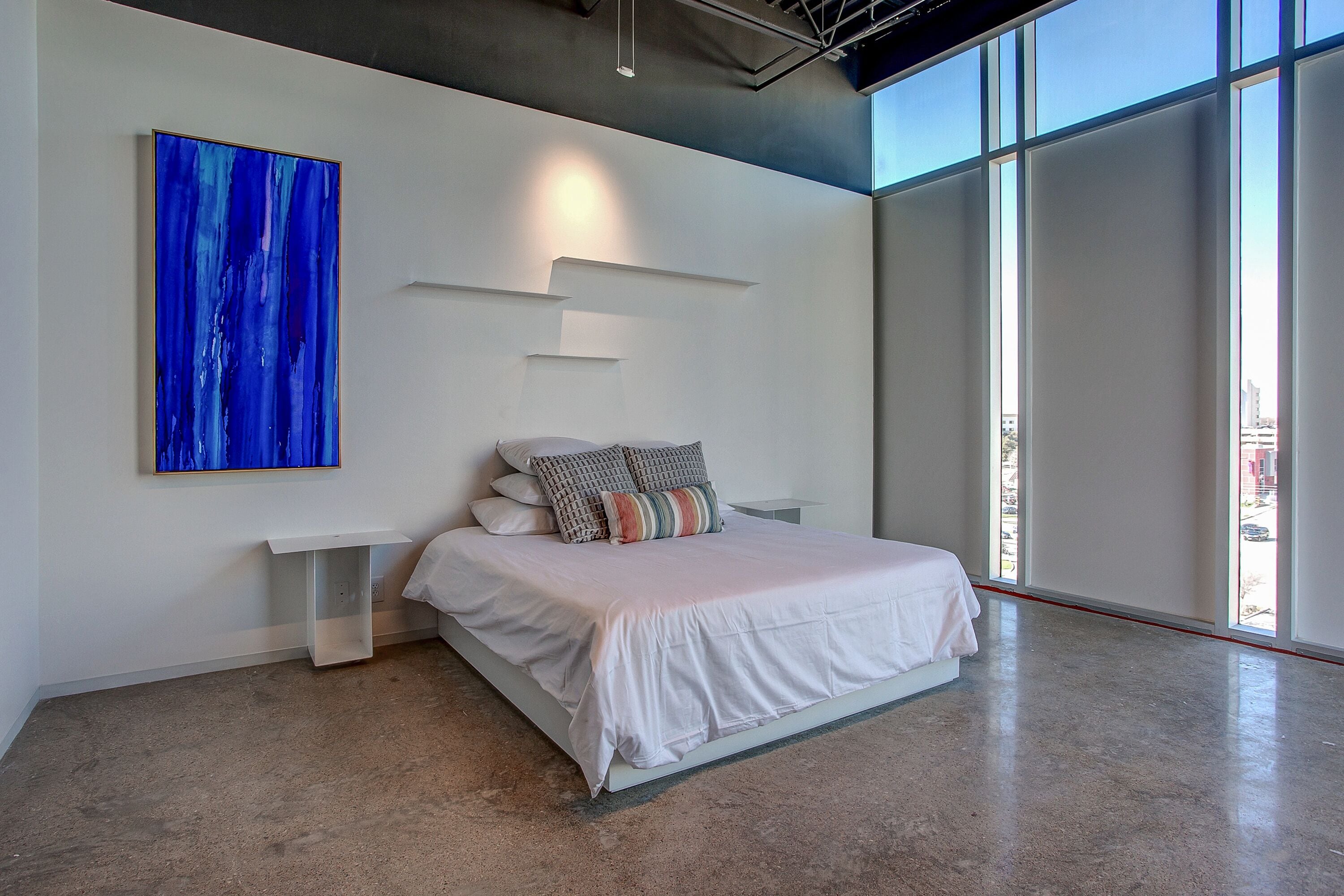 A modern bedroom with a large bed, a white bedside table, a wall-mounted abstract painting, and large windows.