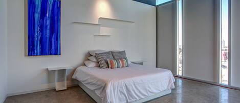 A modern bedroom with a large bed, a white bedside table, a wall-mounted abstract painting, and large windows.