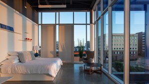 A modern hotel room with a large bed, a small table, and a chair. There is a large window providing a view of the city. - Hotel Revel (Fort Worth)