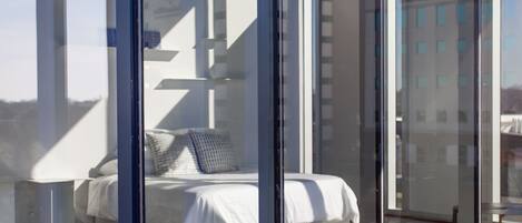 A modern bedroom with a large bed, a bedside table, and a view of a cityscape through floor-to-ceiling windows.