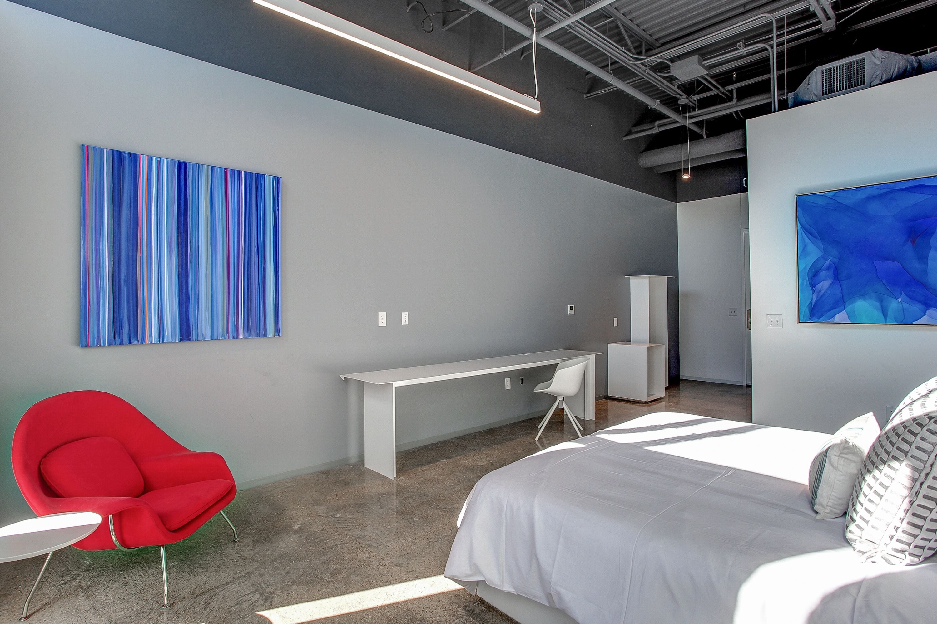 A modern hotel room with a bed, a red chair, a white desk, and two abstract paintings on the walls.