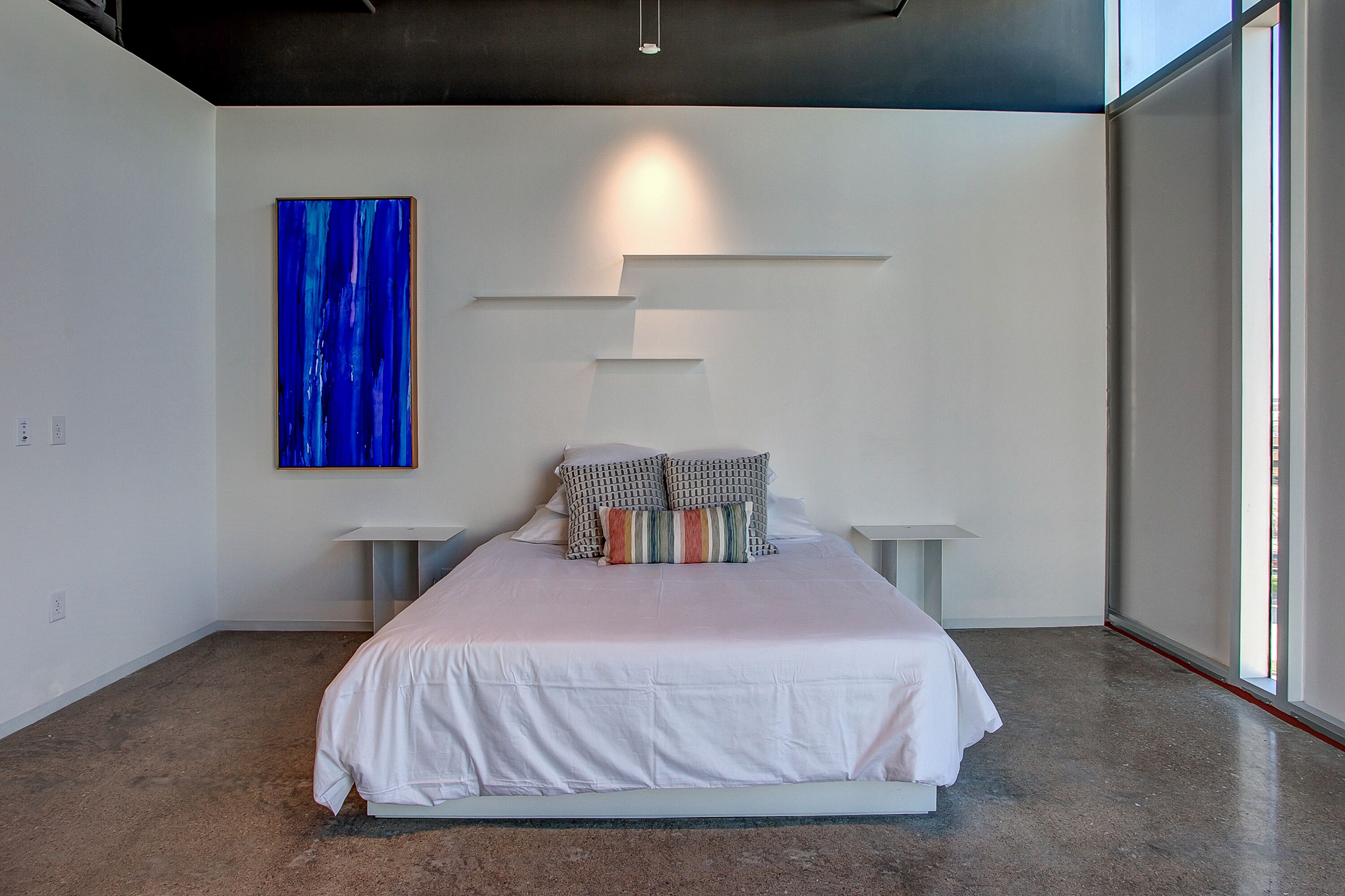 A modern bedroom with a large bed, a blue abstract painting, and a minimalist design.