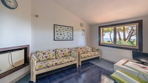 A living room with a sofa, a television, a framed picture of birds, and a window with a view of trees. - Cosy Holiday Apartment "Villetta Vista Mare" with Wi-Fi, Balcony and Terrace (Santa Margherita di Pula)