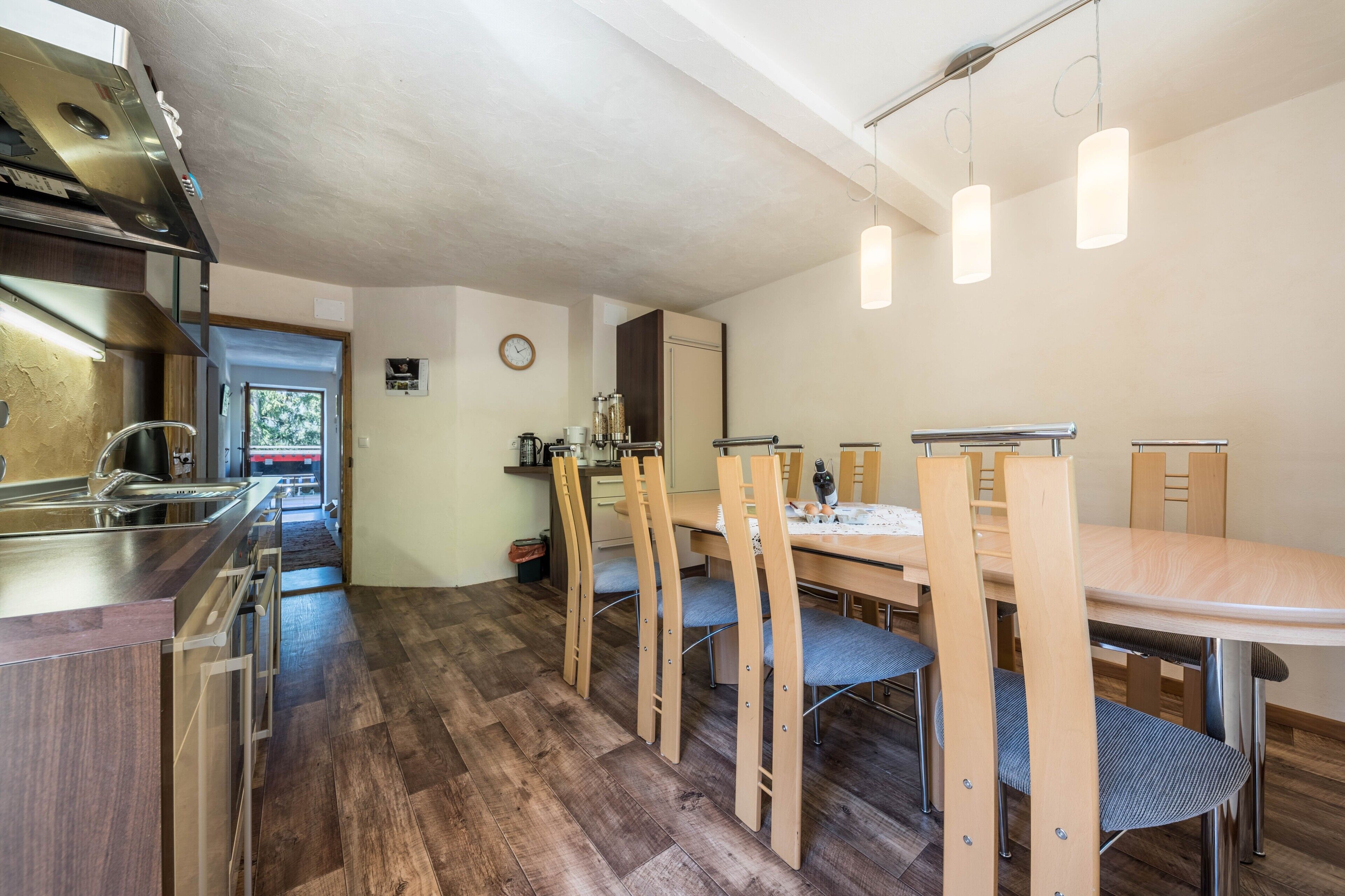 Une cuisine moderne dotée d’une hotte en acier inoxydable, d’une table à manger en bois avec des chaises, et offrant une vue sur l’extérieur par la porte.