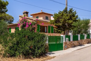 Une maison à deux étages, dotée d’un mur en pierre et de volets verts, entourée de verdure et sous un ciel dégagé.