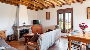 A room with a fireplace, wooden ceiling beams, a television, a dining table with chairs, and a window with a view. - In a quiet location with sea view - El Mirador de Castilnovo (El Palmar (Cádiz))