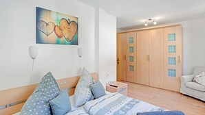 Ein Schlafzimmer mit einem Bett, Kissen, einem Nachttisch, einem Wandbild, einem Kleiderschrank und einem Sessel.