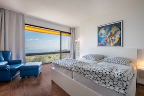 Ein modernes Schlafzimmer mit einem großen Bett, einem blauen Sessel, einem Balkon mit Aussicht und einem farbenfrohen abstrakten Gemälde.