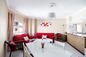 A living room with a glass table, red sofa, wicker chairs, and a kitchen area with a counter. - Family Apartment "Goya 1º" in a Quiet Location (Conil de la Frontera)