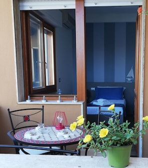 A balcony with a table, chairs, a potted plant, and a bed with a sailboat decoration.