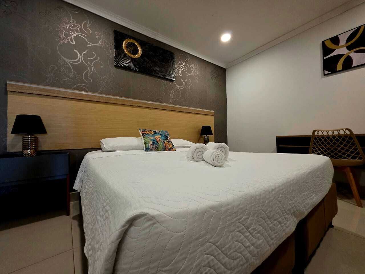 Un lit bien fait, recouvert d’une couette blanche, avec deux serviettes enroulées et un coussin coloré. On y trouve une tête de lit en bois, une horloge murale et une petite table avec une lampe.