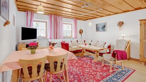 A cozy room with wooden ceiling beams, a dining table with chairs, a sofa, a television, and a window with curtains.