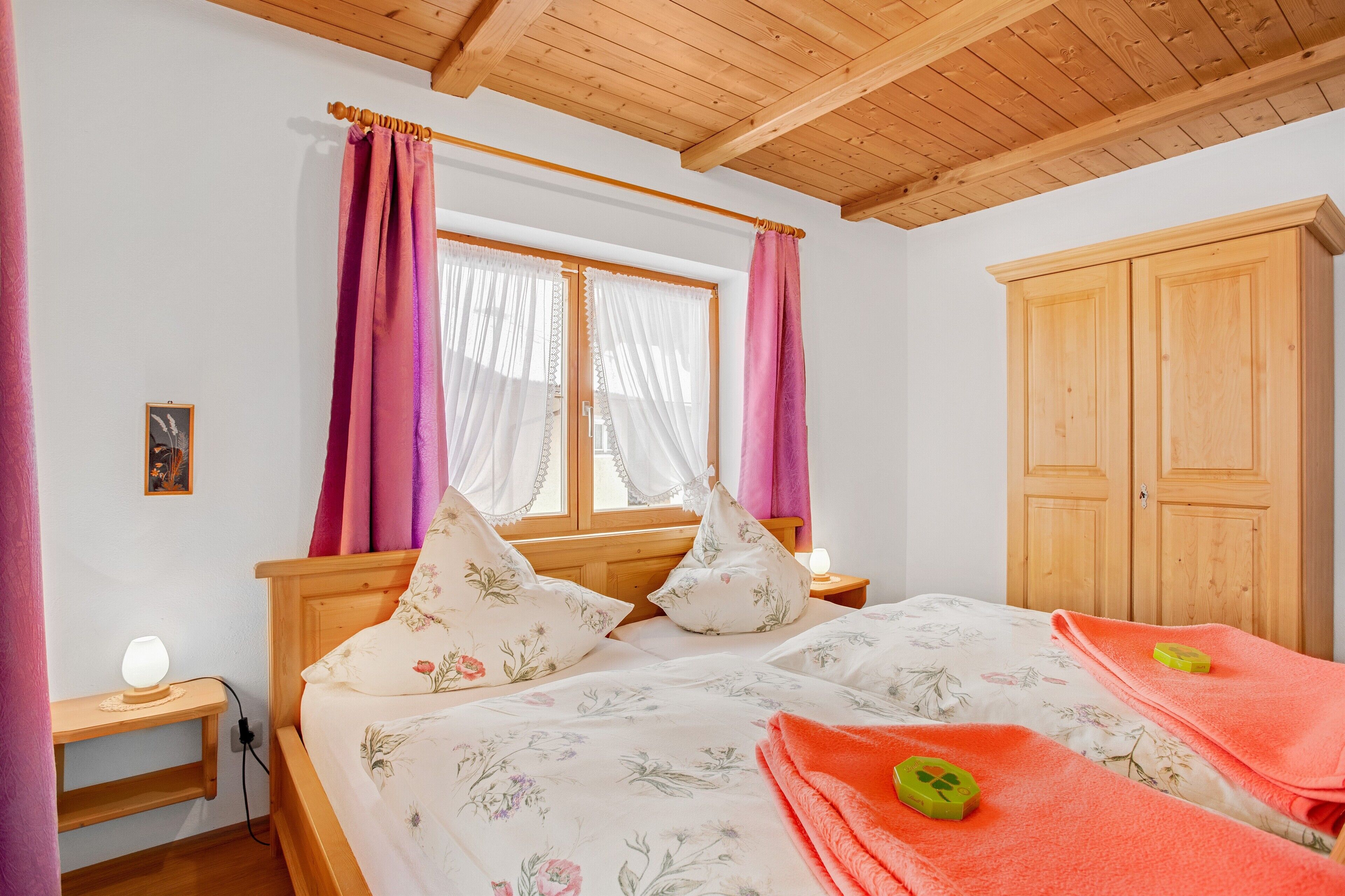 Un dormitorio con techo de madera, cama con ropa de cama floral, mesita de noche con lámpara y armario.