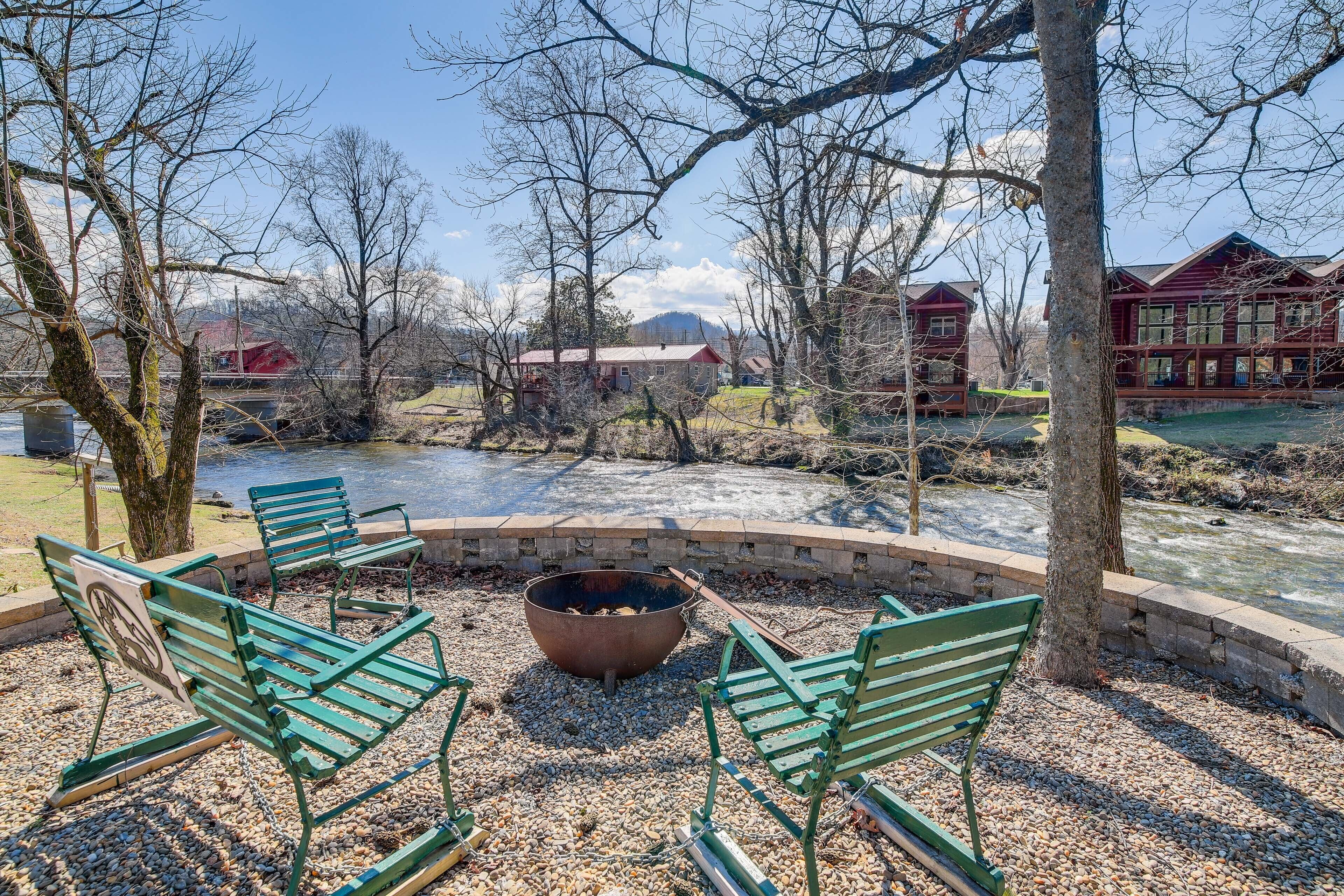 A riverside seating area with benches and a fire pit, surrounded by trees and a view of a red building across the river.