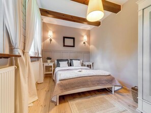 Ein Schlafzimmer mit Holzbalkendecke, einem Bett mit weißer und grauer Bettdecke, zwei Nachttischen mit Lampen, einem Heizkörper und einem Fenster mit Vorhängen.