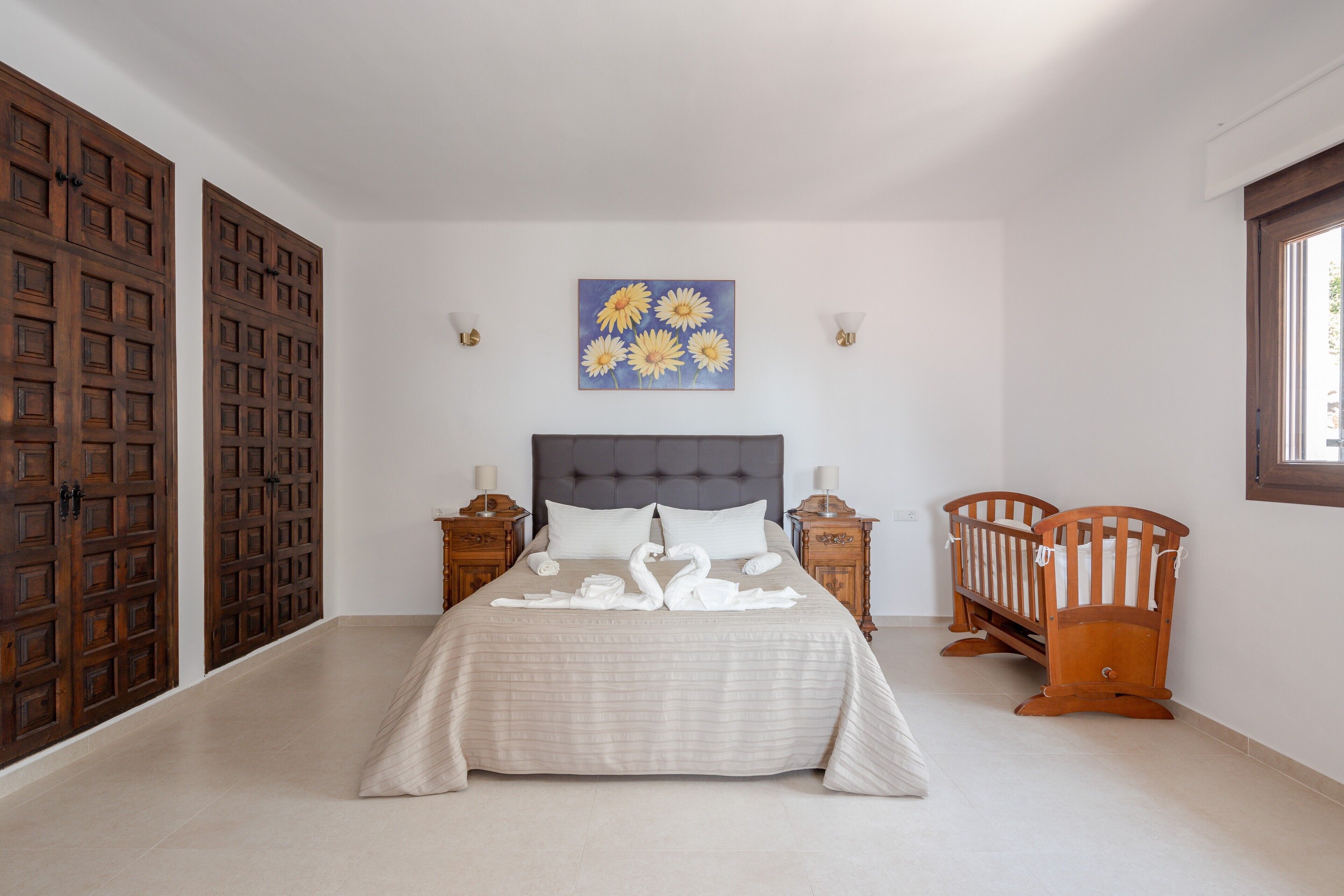 A bedroom with a bed, two nightstands, a crib, a wall-mounted painting, and wooden doors.