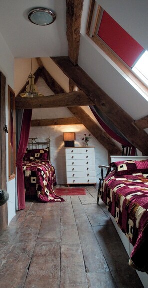 A bedroom with a sloped ceiling, wooden beams, a bed with a patterned bedspread, a dresser, and a window with red curtains.