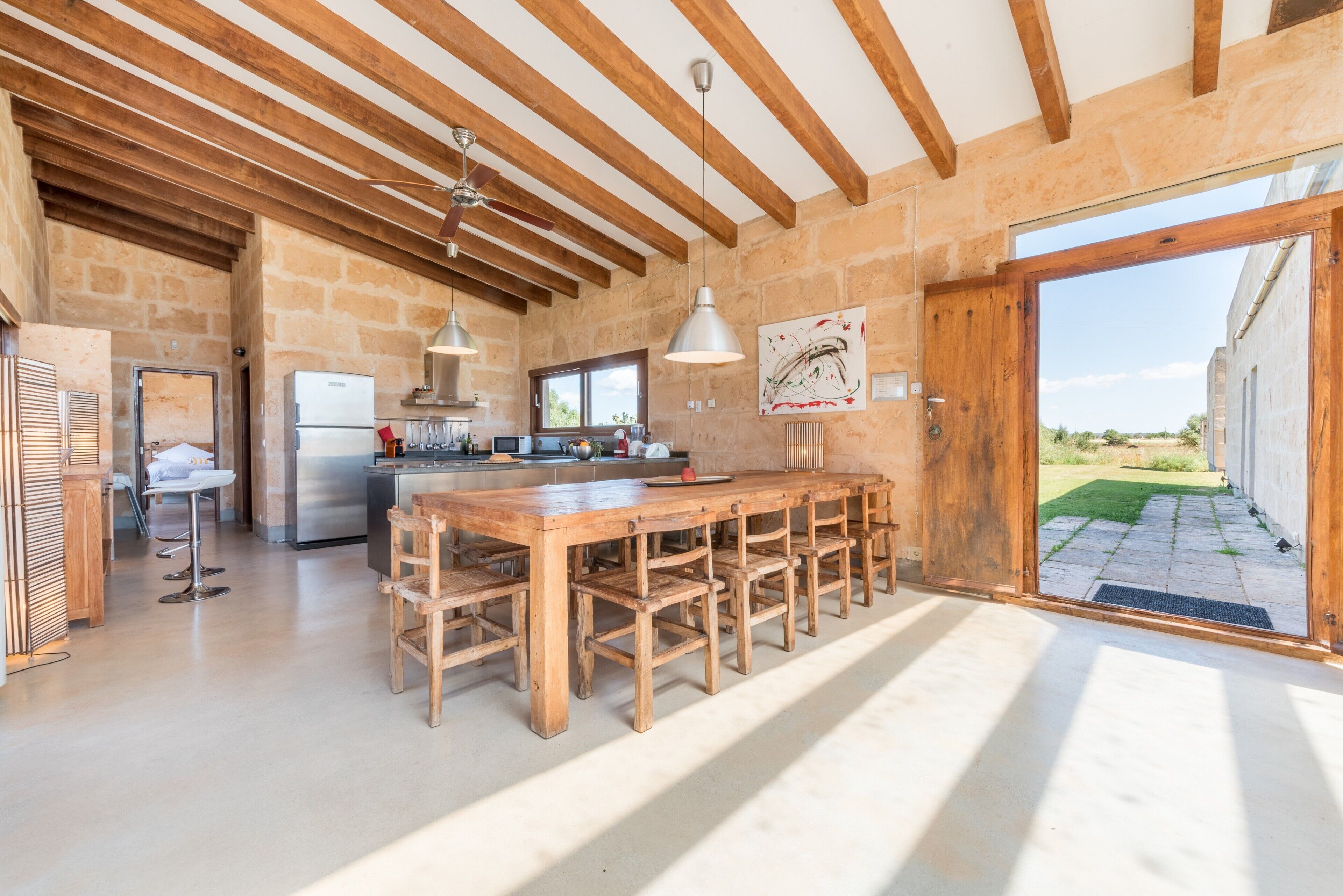 A spacious kitchen with a long wooden table, a built-in refrigerator, and a stone wall.