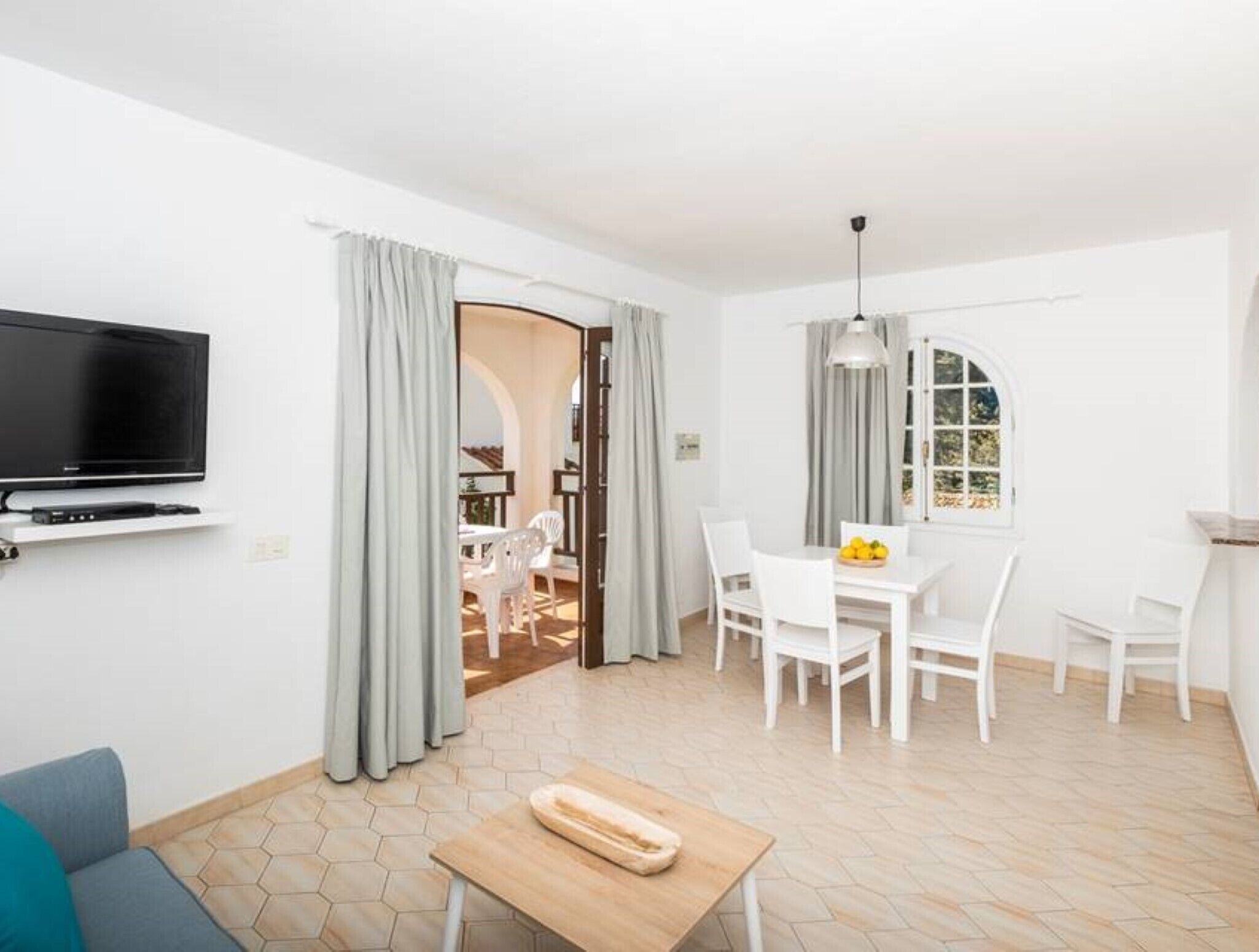 A living room with a flat-screen TV, a dining area with white chairs and a table, and a kitchen area with a window.