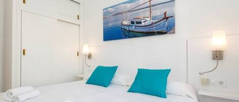 A hotel room with a bed, two pillows, a painting of a boat, and two wall-mounted lamps.