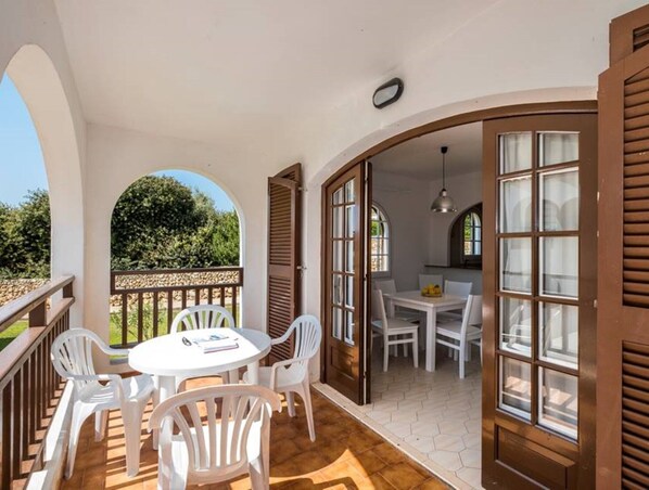Un área de comedor al aire libre, techada, con sillas y una mesa de plástico blanco, comunicada con una sala que tiene una mesa y sillas de comedor.