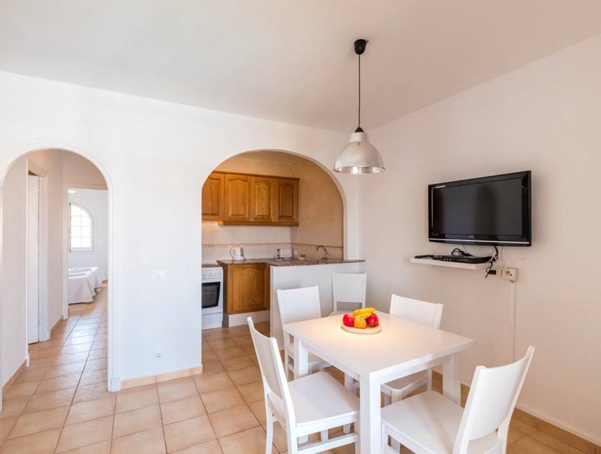 Une cuisine moderne avec des meubles blancs, un coin repas avec une table et des chaises blanches, et un téléviseur fixé au mur.