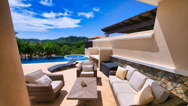 A sunlit outdoor seating area with wicker furniture, a stone-tiled floor, and a view of a swimming pool and lush greenery. - Luxury Villa in prestigious Pristine Bay, home of the Black Pearl Golf Course (French Harbor)