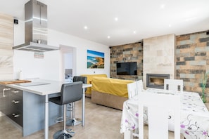 A modern kitchen with a stainless steel range hood, a dining area with a table and chairs, a sofa with a yellow cover, and a fireplace.