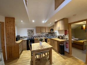 A modern kitchen with wooden cabinets, a dining table, and a small island. There is a sink, stove, and refrigerator. The kitchen has a tiled backsplash and wooden flooring. - Countryside Cottage in the Mendip Hills, with garden (Cross, Axbridge, Somerset)