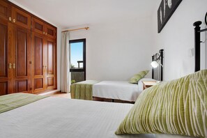 Une chambre avec deux lits, une armoire en bois, une table de chevet avec une lampe, et une vue sur l’extérieur.